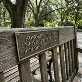 Wooden bench with a bronze plaque reading 'Honoring the past, Enjoying the present, Building the future' in a park setting.