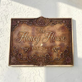 Decorative address sign for 'The Honey House' with floral and bee designs on a white wall.