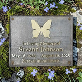 Memorial plaque with butterfly design and engraved text on a natural background with flowers.