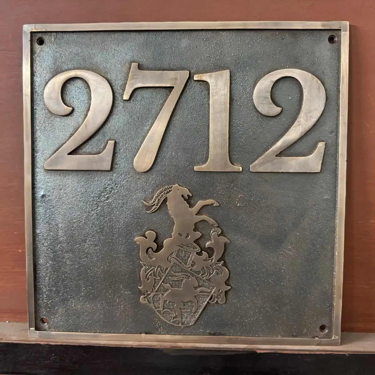 Metallic house number sign with '2712' and a coat of arms on a wooden background
