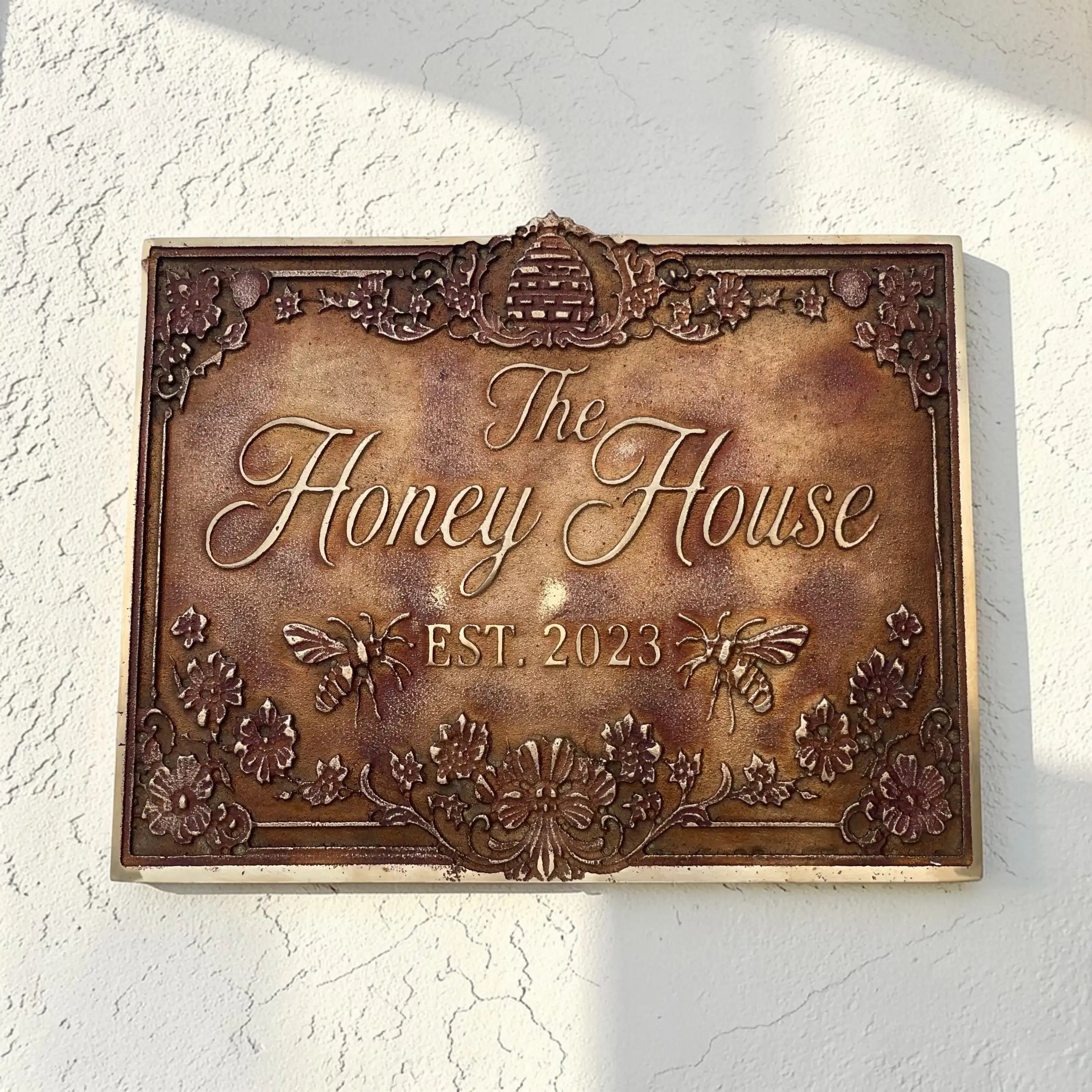 Decorative address sign for 'The Honey House' with floral and bee designs on a white wall.