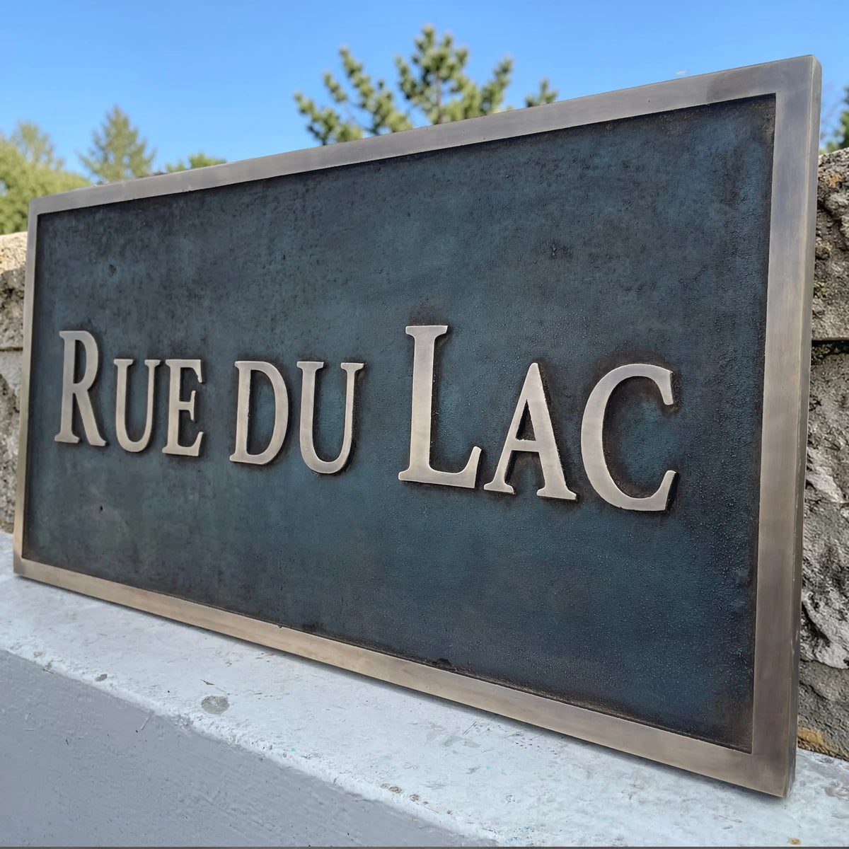 Bronze address sign with 'RUE DU LAC' text against a stone wall and blue sky background