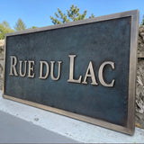 Bronze address sign with 'RUE DU LAC' text against a stone wall and blue sky background