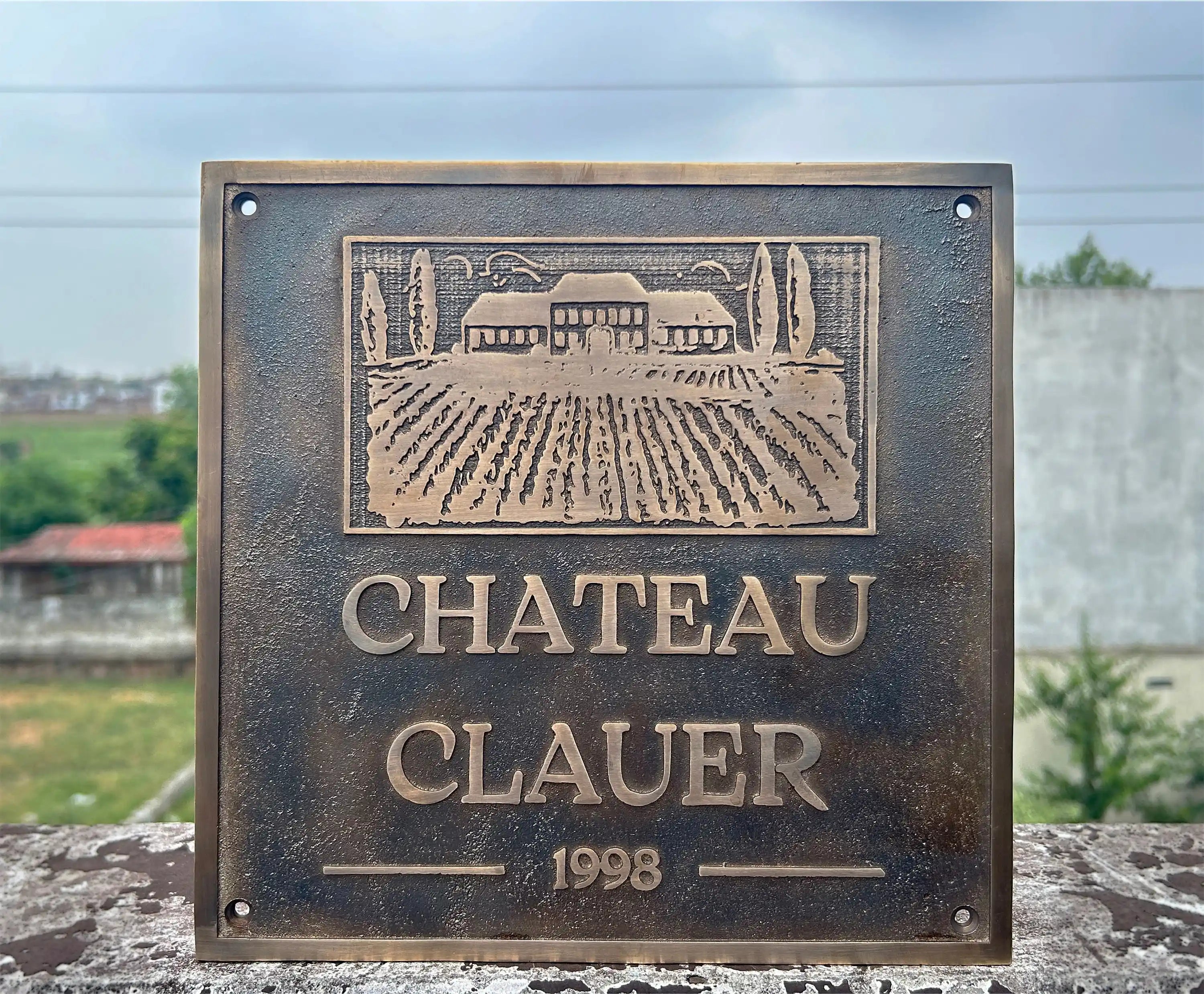 Bronze plaque with 'Chateau Clauser' and year 1998 on a stone surface with a vineyard background.