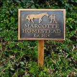 Metal sign with horse and person silhouette, 'Marcotte Homestead Est. 2024', on a wooden stake against green foliage.